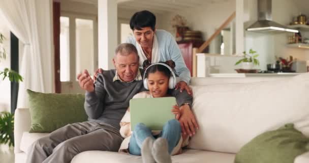 Feliz, falando avós e criança com um tablet para jogos, um desenho animado ou filme em uma sala de estar em casa. Sorriso, família e um homem idoso, mulher e criança com tecnologia e fones de ouvido para a educação. - Filmagem, Vídeo