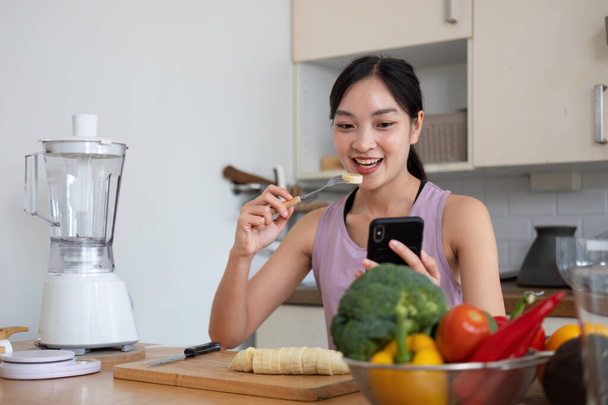 woman with casual clothes making salad and using mobile to count calories and plan diet. Happy woman consulting recipe instructions on smartphone while chopping vegetables in modern kitchen. - Foto, Bild