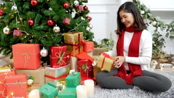 Mujer asiática desenvolver la caja de regalo de Navidad bajo el árbol de Navidad consiguió regalo regalo sorpresa. Las mujeres asiáticas unbox caja de regalo sentado bajo la decoración bauble bola árbol de Navidad fiesta de Navidad invierno celebrar en la sala de estar - Metraje, vídeo