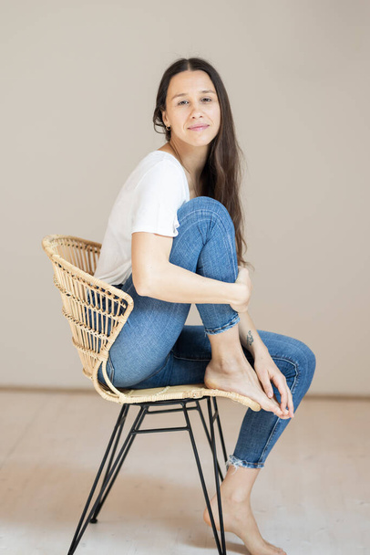 Retrato de mulher bonita confiante com cabelos castanhos longos, vestindo roupas casuais, sentado na cadeira em jeans apertados e t-shirt branca, fundo de estúdio - Foto, Imagem