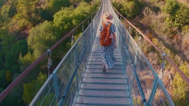 Kobieta turystka przechodzi przez most przez rzekę. Młoda kobieta spaceruje mostem wiszącym przez rzekę przed jesiennym lasem. Dziewczyna w kapeluszu. - Materiał filmowy, wideo