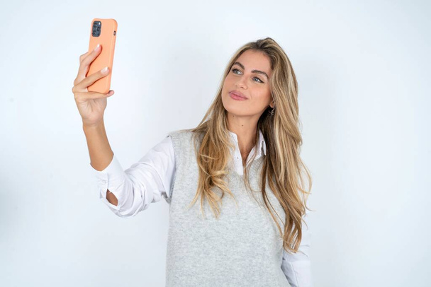 mujer caucásica vistiendo camisa blanca sonriendo y tomando una selfie lista para publicarla en sus redes sociales. - Foto, imagen