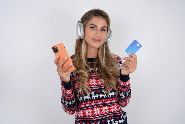 Kaukasische vrouw draagt kerst trui draagt stereo koptelefoon op oren houdt moderne mobiele telefoon en creditcard - Foto, afbeelding