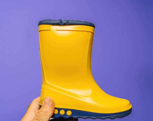 POV: Male hand presents vibrant yellow childrens boots against a white and violet backdrop - Фото, изображение