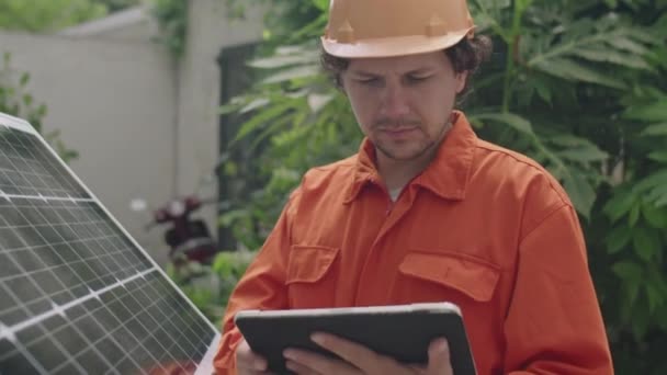 Keskipitkä laukaus ammatillinen PV asentaja työvaatteet ja kypärä seisoo aurinkopaneeli ulkona ja digitaalinen tabletti - Materiaali, video