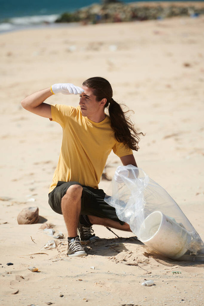Junger Mann trägt Handschuhe beim Müllsammeln am Strand in Plastiktüte - Foto, Bild