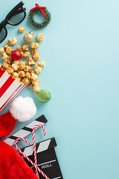 Ταινία με θέμα τα Χριστούγεννα με εορταστικές λιχουδιές. Top view κάθετη φωτογραφία εορταστική ταινία clapper, 3D γυαλιά, γλυκά μπαστούνια καραμέλα, καπέλο του Άη Βασίλη, στολίδια, μικρό στεφάνι, ποπ κορν σε χιονισμένο φόντο - Φωτογραφία, εικόνα