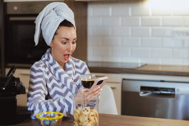 Atrakcyjna kobieta w szlafroku używa smartfona podczas porannej kawy w domu. - Zdjęcie, obraz