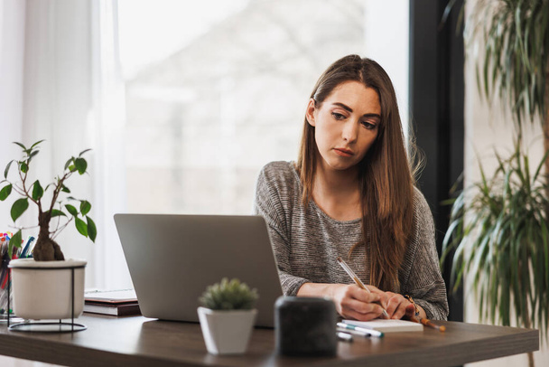 Jeune femme entrepreneure regardant réfléchi tout en travaillant à partir de son bureau à domicile. - Photo, image