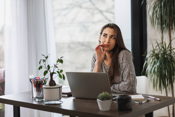 Criativa jovem mulher olhando pensativo enquanto trabalhava no laptop de seu escritório em casa. - Foto, Imagem