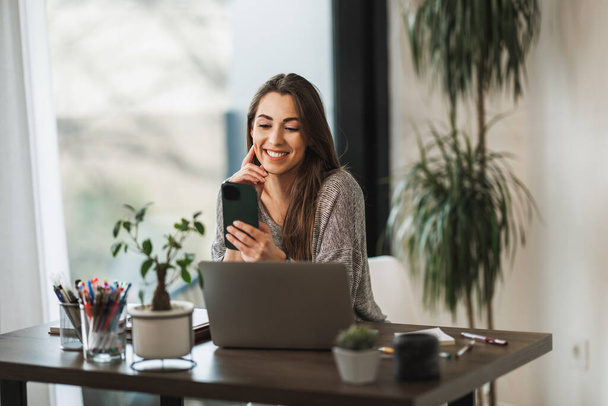 Foto van een jonge glimlachende vrouw met behulp van een mobiele telefoon en laptop tijdens het werken vanuit huis. - Foto, afbeelding