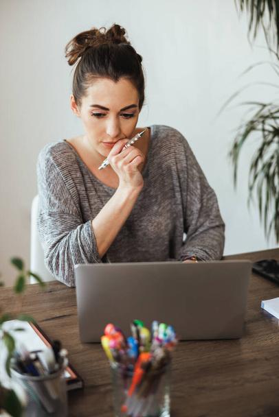Jovem designer mulher olhando pensativo enquanto trabalhava no laptop de seu escritório em casa. - Foto, Imagem