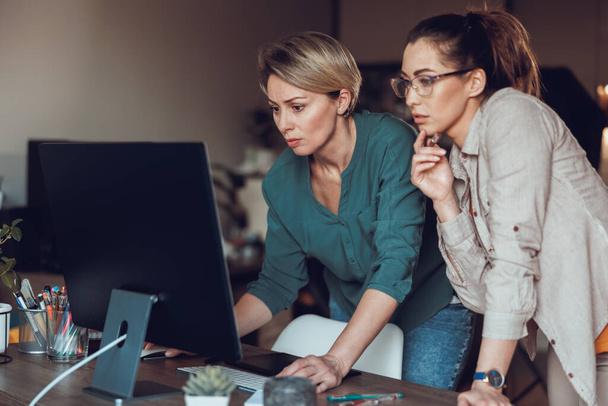 Two female entrepreneur looking thoughtful while working together on computer, doing project from home office. - Photo, Image