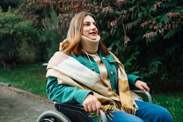 Dışarıda tekerlekli sandalyede oturan 30 'lu yaşlarda boyunluk takan bir kadın. - Fotoğraf, Görsel