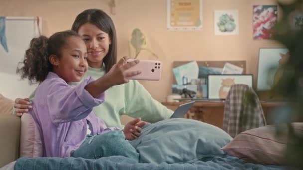Happy 10 year old African American girl taking selfie portrait on smartphone with mom sitting together on bed in cozy kids bedroom with peach walls - Footage, Video