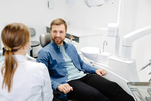 Ξανθιά γυναίκα οδοντίατρος στο οδοντιατρείο μιλάει με τον άνδρα ασθενή και ετοιμάζεται για θεραπεία. Όμορφος γενειοφόρος στην καρέκλα του οδοντιάτρου κοιτάζει το γιατρό του με χαμόγελο. - Φωτογραφία, εικόνα