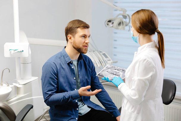 Jonge aantrekkelijke man die tandarts bezoekt, zit in een tandartsstoel in een moderne lichtkliniek. Jonge vrouw tandarts holding x ray afbeelding - Foto, afbeelding