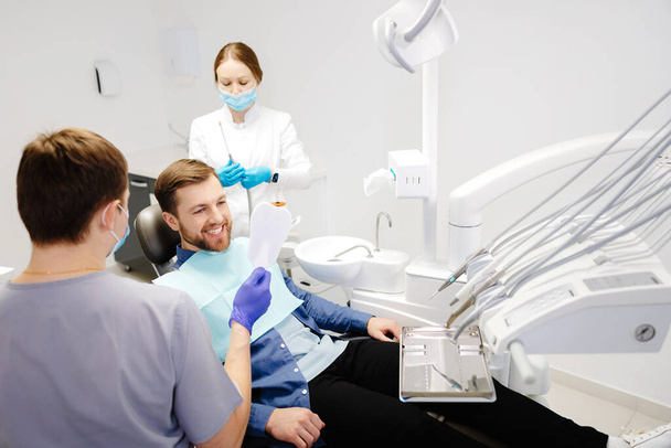 Hombre guapo paciente mirando su hermosa sonrisa sentado en el consultorio dental. - Foto, Imagen