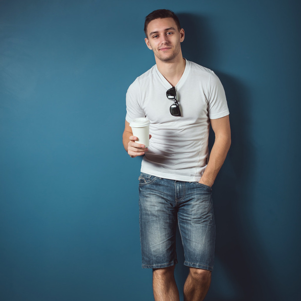 Молодой привлекательный красивый мужчина в джинсах в белых футболках в джинсах, стоящий у стены и улыбающийся, держа кофе
. - Фото, изображение
