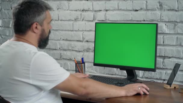 Homem de meia-idade barbudo olha para a tela verde do computador e escreve algo no caderno. Homem trabalha no escritório moderno com parede de tijolo branco - Filmagem, Vídeo