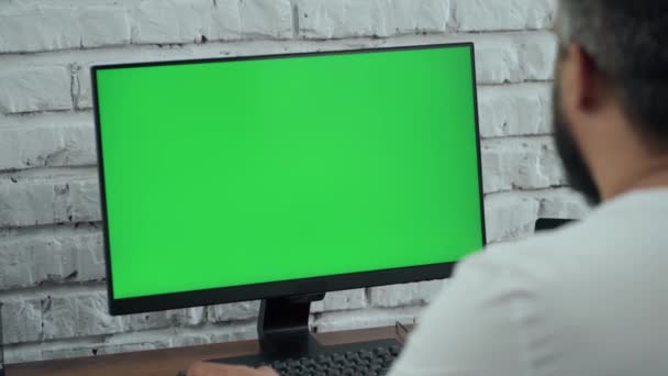 Бородатый мужчина средних лет работает за настольным компьютером с зеленым макетом экрана. Интерьер - современный офис с белой кирпичной стеной - Кадры, видео