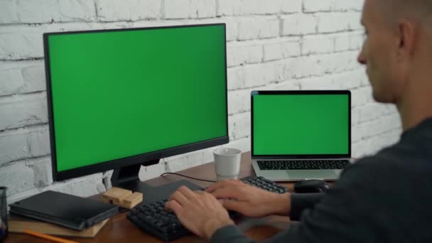 El hombre trabaja en la computadora de escritorio y el ordenador portátil con pantallas verdes en la oficina. Tecla de croma en la computadora y la pantalla del ordenador portátil - Imágenes, Vídeo