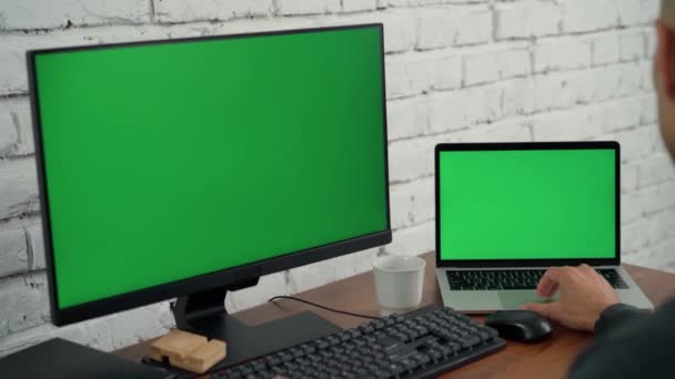 Mies työskentelee pöytätietokoneella ja kannettavalla tietokoneella, jossa on vihreät näytöt toimistossa. Chroma avain tietokoneen ja kannettavan tietokoneen näyttö - Materiaali, video