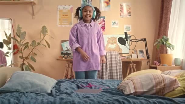 Μέτριο πλάνο του χαρούμενου African American gen z κορίτσι φορώντας ασύρματα ακουστικά διασκεδάζοντας ενώ χορεύουν σε ζωντανή παιδικό υπνοδωμάτιο - Πλάνα, βίντεο