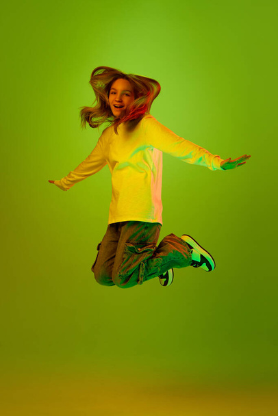 Ein hübsches Kind, Mädchen lässig gekleidet, hüpft fröhlich davon und genießt vor grünem Studiohintergrund in gemischtem Farblicht. Konzept von Glück, Gesichtsausdruck, Mensch, Emotionen, Kindheit, Einkaufen. - Foto, Bild