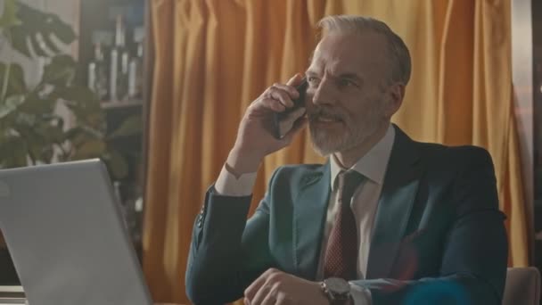 Elegant mature Caucasian businessman in formalwear making phone call to business partner and looking at wristwatch while working at table in restaurant - Footage, Video