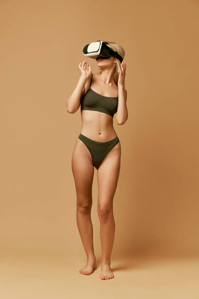 Full length foto van mooie slanke vrouw in ondergoed, lingerie en VR bril poseren tegen pastel beige studio achtergrond. Concept natuurlijke schoonheid, gezondheid en lichaamsverzorging, anti-cellulitis, vrouwelijkheid. - Foto, afbeelding