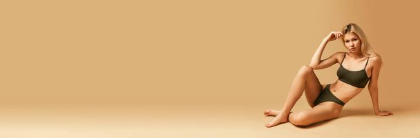 Баннер. Флайер. Стройная, стройная леди в оливковом белье, лежащая на полу изолированная на фоне пастельных, бежевых студий. Фитнес. Концепция красоты, ухода за телом, здоровья, диеты, косметики, спа. Копирование места для рекламы. - Фото, изображение