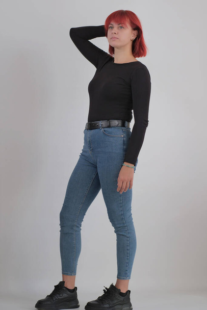 Una giovane donna attraente con una breve acconciatura arancione in una t-shirt nera e jeans in posa in studio su uno sfondo bianco. - Foto, immagini
