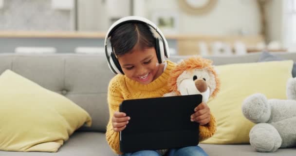 Tablet, Kopfhörer und glückliches Mädchen oder Kind beim Lernen, zu Hause oder beim Hören von Übersetzungen oder Audio auf dem Sofa. Kind mit Teddybär, Spiele oder lustige Komödie oder Zeichentrick auf digitaler Technologie ansehen. - Filmmaterial, Video