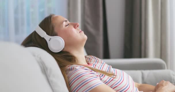 Fiatal nő pihentető kényelmes kanapé csukott szemmel visel fejhallgató, lány élvezi hallgatni a zenét és a hang pihentető otthon. Kiváló minőségű 4k felvételek - Felvétel, videó