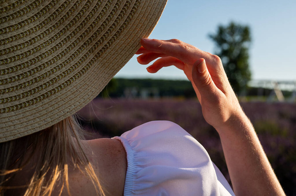 Ξανθό μακρυμάλλη κορίτσι σε ένα πεδίο λεβάντα στο ηλιοβασίλεμα σε ένα ψάθινο καπέλο ισιώνει με το χέρι της και ένα λευκό φόρεμα. Πίσω όψη - Φωτογραφία, εικόνα