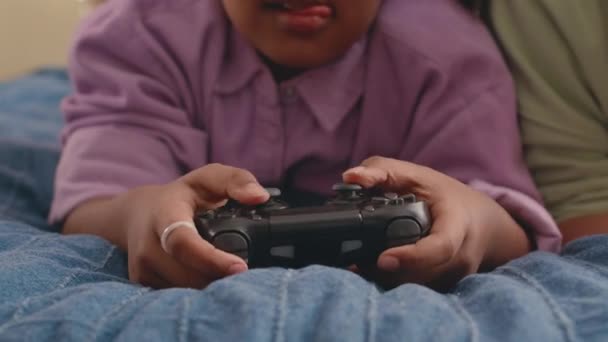 Medio primer plano de chica afroamericana usando controlador mientras juega videojuegos con mamá en la cama en casa - Imágenes, Vídeo