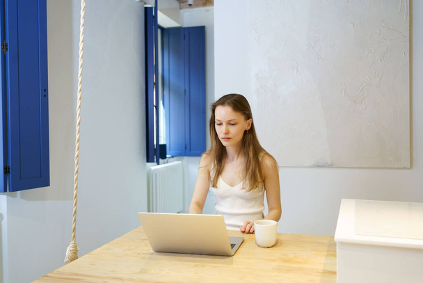 Een jonge vrouw typt op een laptop terwijl ze aan tafel zit in de woonkamer. Het concept van afstandsonderwijs en -coaching, online cursussen en lessen - Foto, afbeelding