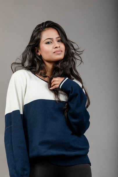 Ινδή νεαρή γυναίκα μοντέλο σε casual winterwear σε γκρι φόντο. Μακριά μαύρα μαλλιά μοντέλο φορώντας μαύρα κολάν, μπλε και άσπρο φούτερ. - Φωτογραφία, εικόνα