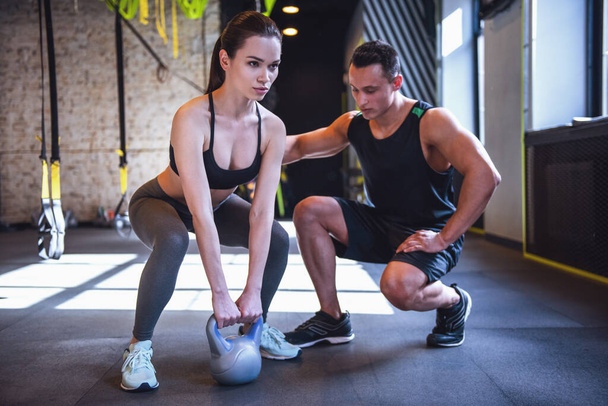 Beautiful sports girl is doing exercises with a kettlebell while working out with a personal trainer in gym - Photo, Image