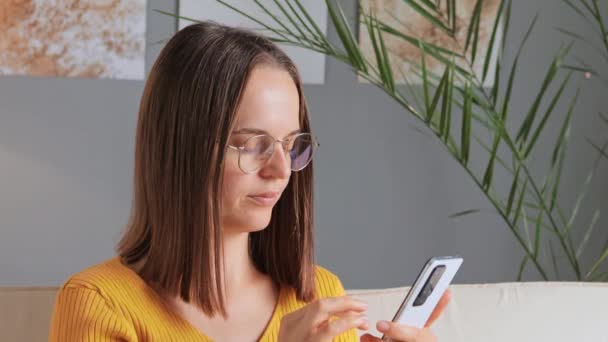 Tired beautiful woman taking off glasses while using mobile phone long hours feeling pain in her eyes rubbing eye sitting at home interior in living room browsing internet pages scrolling online. - Video, Çekim