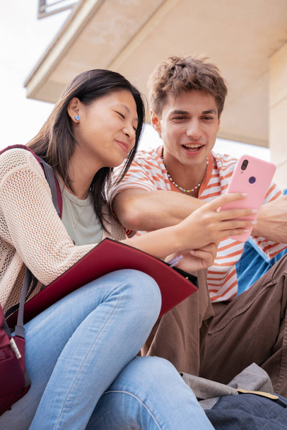 Gülümseyen çok ırklı öğrenciler dışarıda oturmuş internette sohbet etmek için cep telefonu kullanıyorlar. Üniversite yaşam konsepti - Fotoğraf, Görsel
