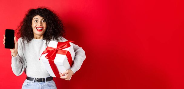 Ημέρα του Αγίου Βαλεντίνου και εραστών. Ενθουσιασμένη χαμογελαστή γυναίκα με σγουρά σκούρα μαλλιά, που δείχνει το smartphone άδειο οθόνη και κρατώντας δώρο έκπληξη στις διακοπές, που δείχνει online promo, κόκκινο φόντο. - Φωτογραφία, εικόνα