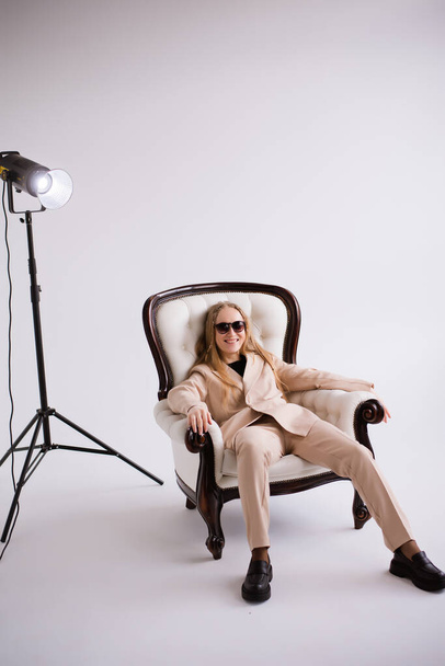 Nő, modell, blogger, szőke napszemüvegben, széken ülve a fehér fotóstúdióban, elkülönítve. Formális bézs nadrágkosztümbe öltözve - Fotó, kép