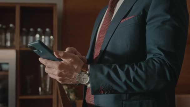 Środkowa część nierozpoznawalnego biznesmena w eleganckim garniturze i drogim zegarku przewijającym się na smartfonie w pomieszczeniu - Materiał filmowy, wideo