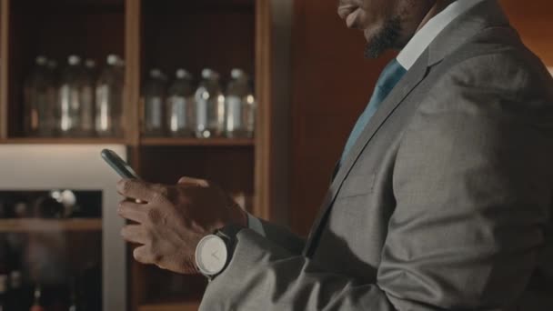 Tilt up shot of successful young African American businessman in elegant grey suit texting on smartphone while standing in modern restaurante with bar - Imágenes, Vídeo