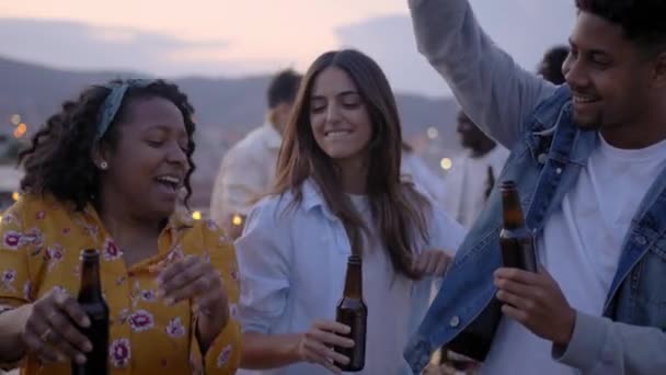 Hidasliikkeinen ryhmä innostuneita monirotuisia nuoria juhlimassa kattobileitä juomassa olutta. Ystäviä tanssimassa ja laulamassa viikonloppuna. Iloinen gen z nauttia yön vapaa ulkona. - Materiaali, video