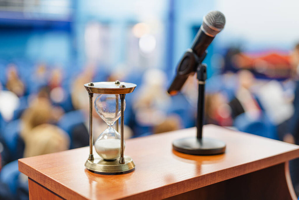 Gros plan d'une horloge de sable à côté d'un microphone dans la salle de conférence pleine de personnes - Photo, image