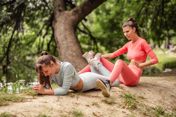Active, fit females doing stretching exercises in a city park. Friends training outdoors, exhibiting energy and stamina. Vibrant urban sport routine among trees. - Фото, изображение