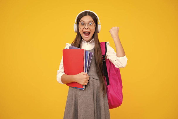 Iskolás lány, 12, 13, 14 éves, fülhallgatóban és elszigetelt stúdió háttérről szóló könyvekben. Iskolások hátizsákkal. Izgatott tinédzser lány - Fotó, kép